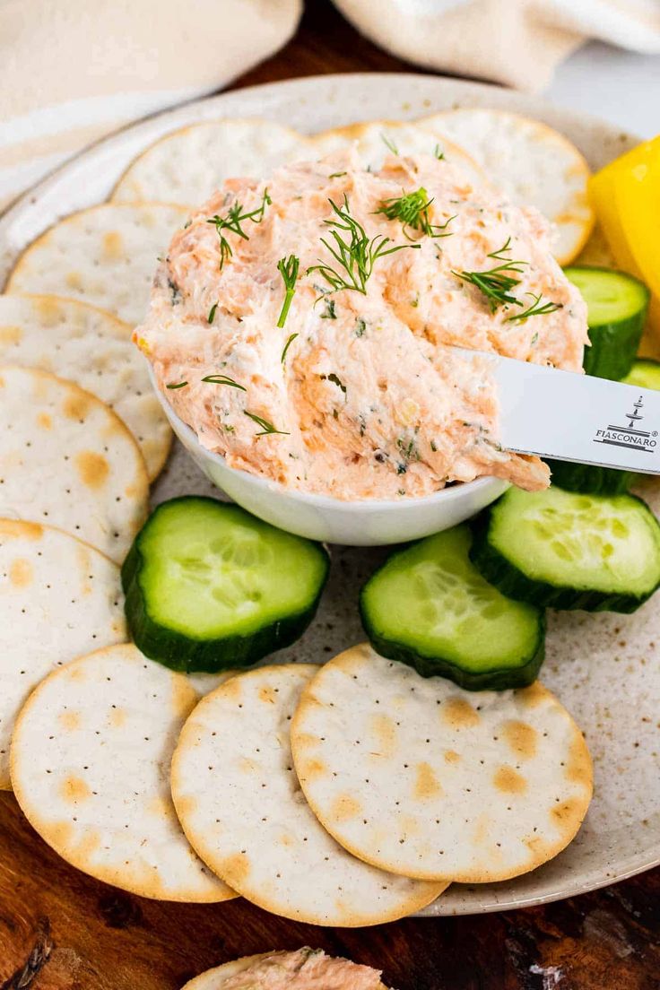 a plate with crackers, cucumber and dip on it next to lemon wedges