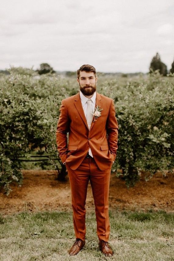 a man in an orange suit and tie standing in front of some bushes with his hands in his pockets