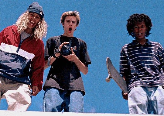 three young men standing on top of a building holding skateboards and looking at the camera