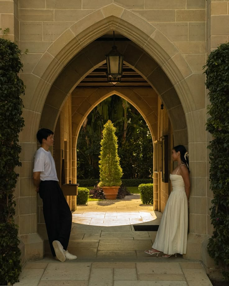 a man and woman standing in front of an archway with plants on either side, looking at each other