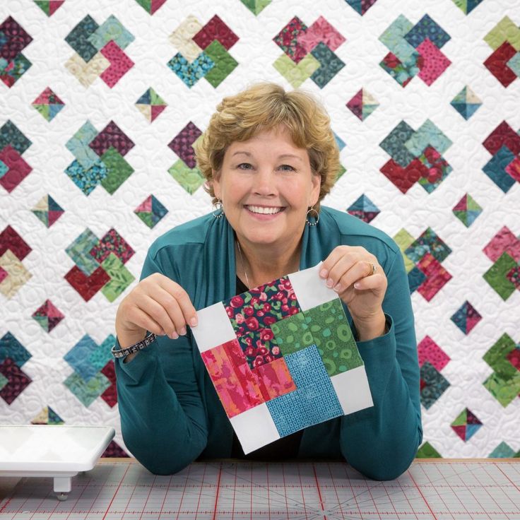 a woman sitting at a table holding up a piece of quilted material with the background made from squares