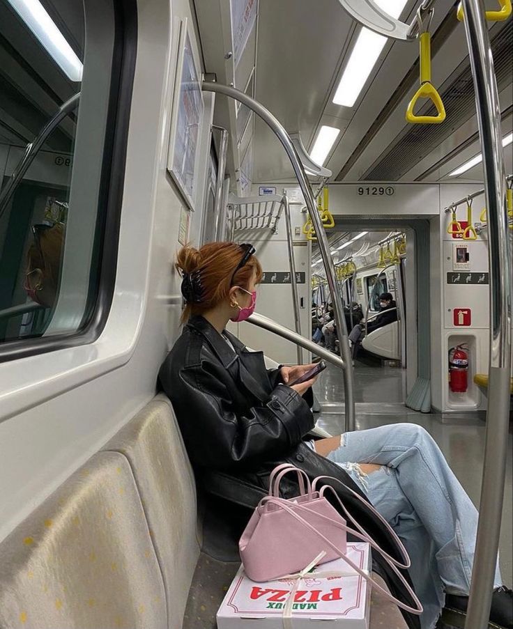 a woman sitting on a subway train looking at her cell phone while wearing a mask