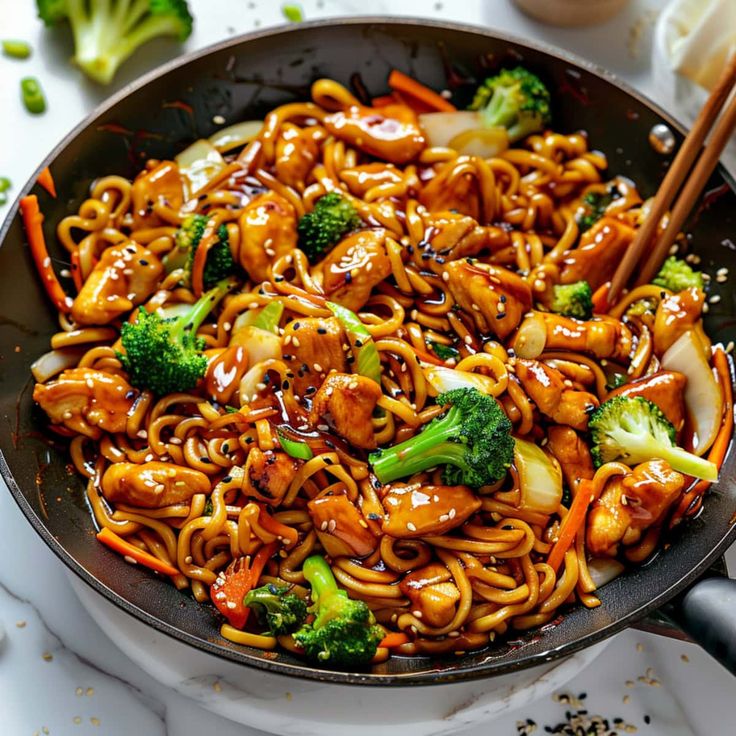 a wok filled with noodles, broccoli and chicken on top of a table