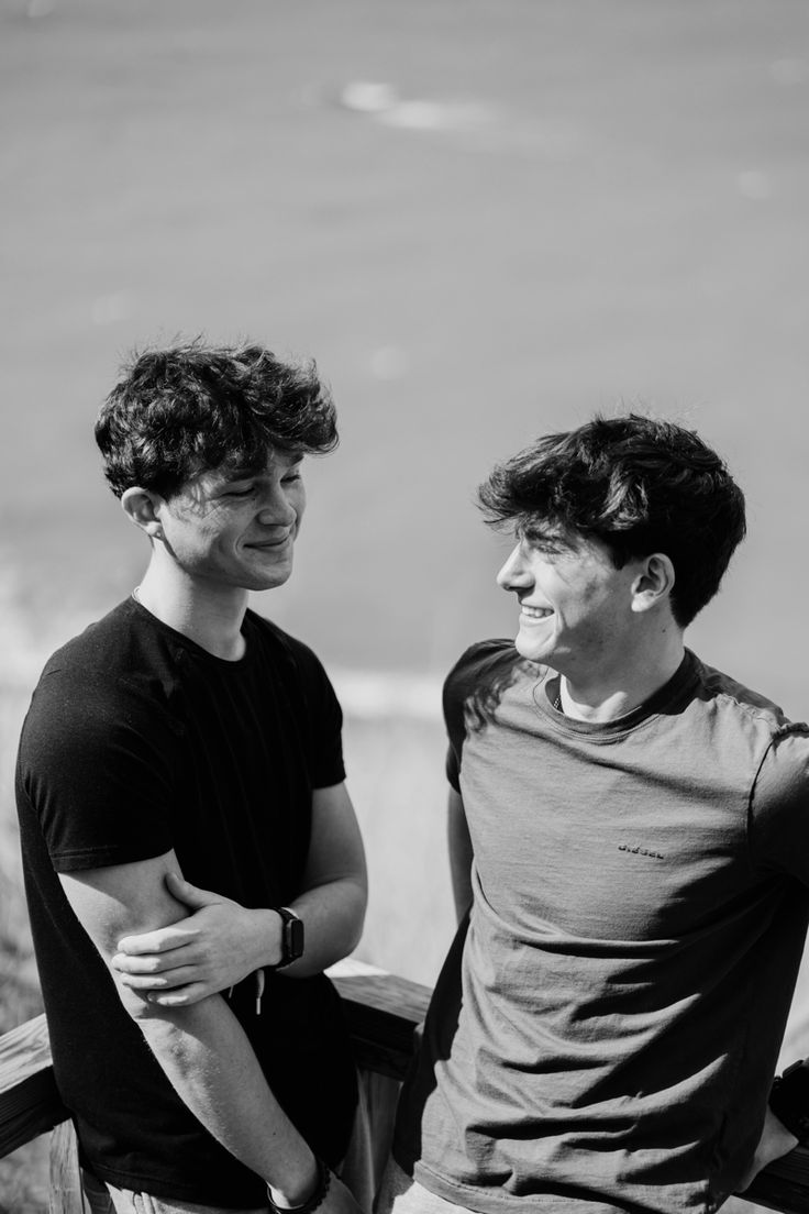 two young men sitting next to each other on a bench looking at the ocean in black and white