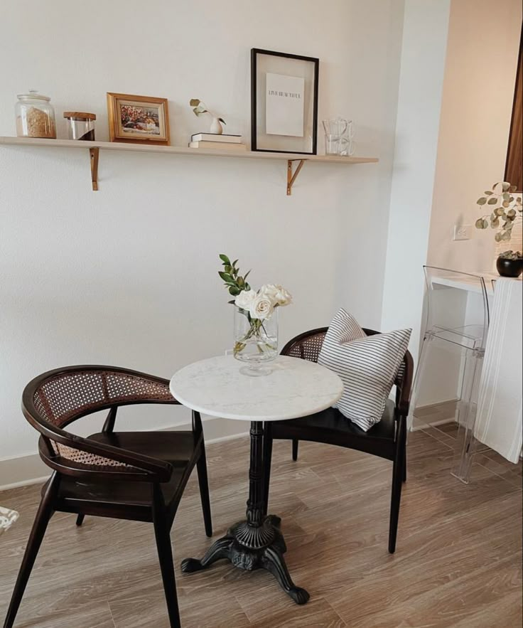 two chairs and a table in a room with white walls, wood flooring and shelves