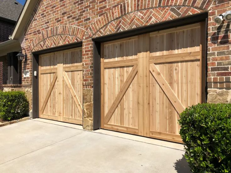 two wooden garage doors in front of a brick building with bushes and shrubs around them