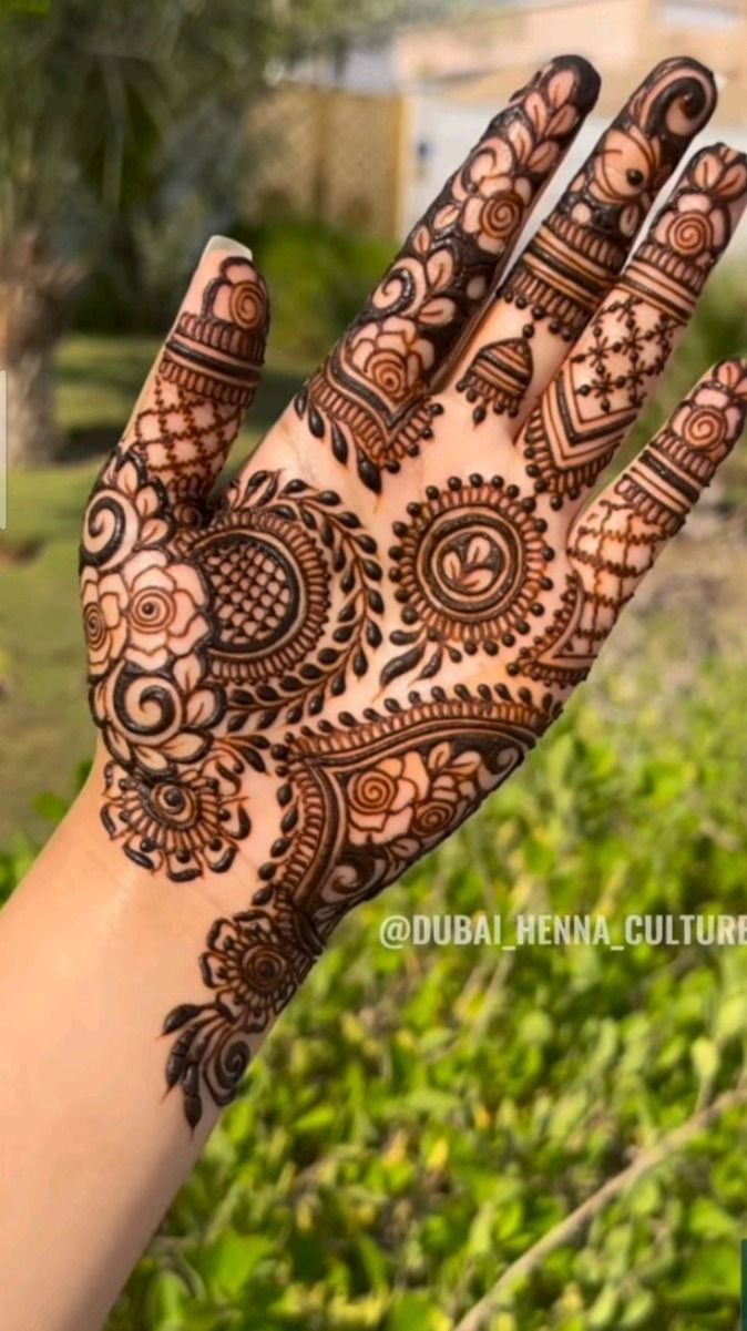 a hand with henna tattoos on it and green plants in the back ground behind it