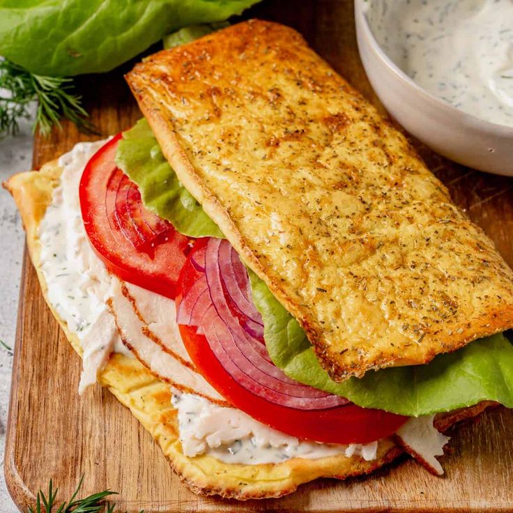 a sandwich with lettuce, tomato and onion on a cutting board next to a bowl of ranch dressing