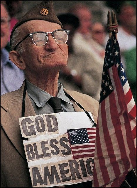 an old man holding a sign that says god blessing america with an american flag in the background