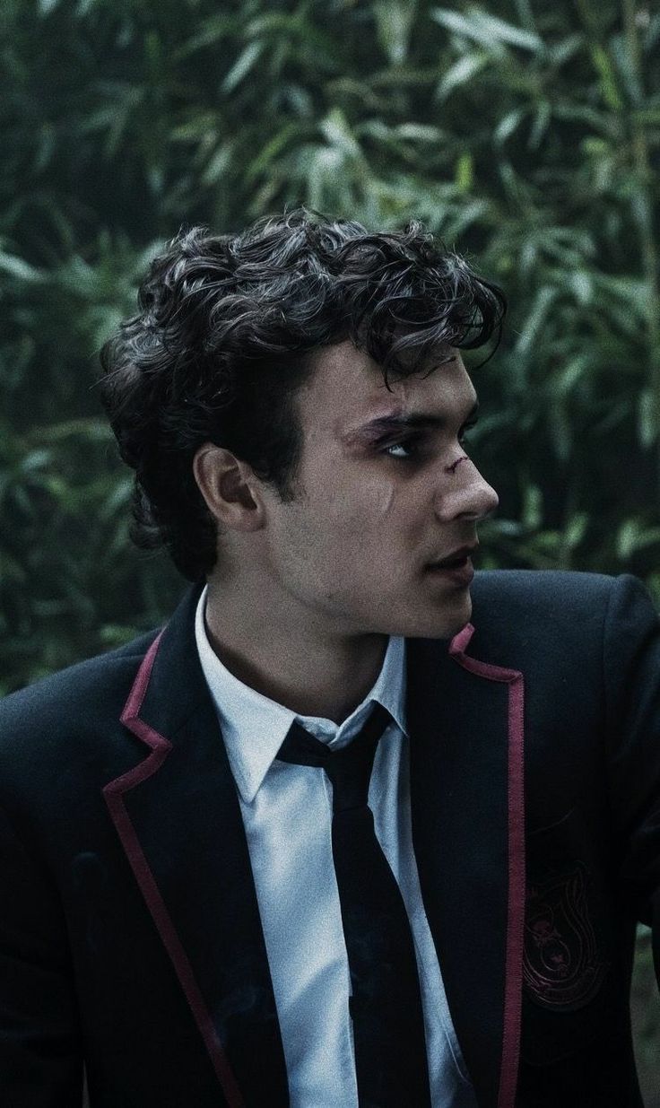 a man in a suit and tie sitting next to a bush looking off into the distance