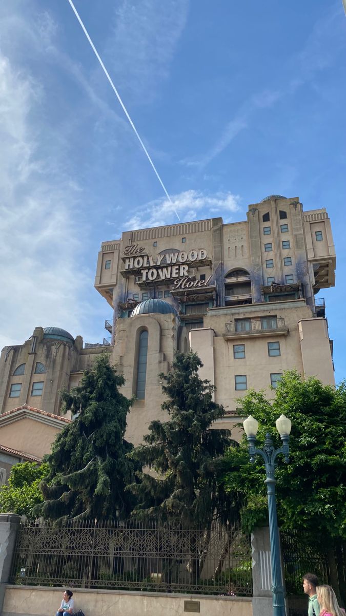 people are sitting on the sidewalk in front of a large building with an airplane flying overhead