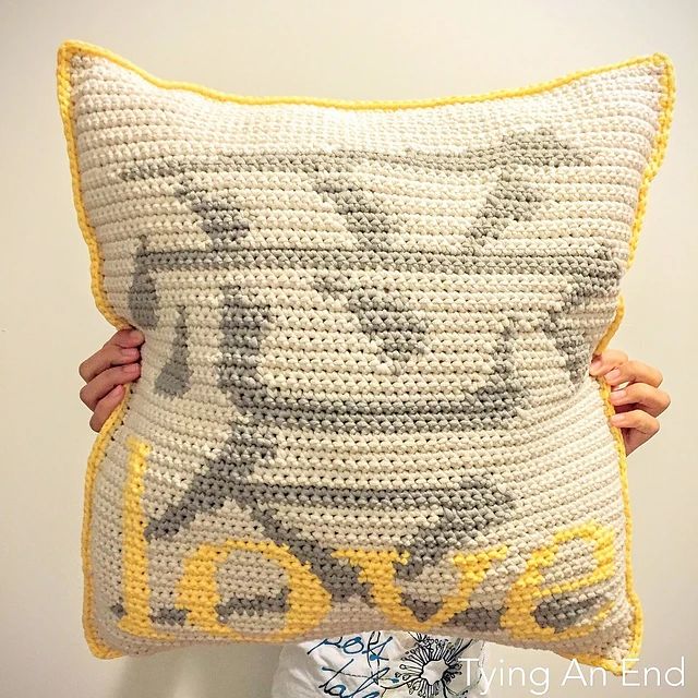 a person holding a crocheted pillow in front of a white wall with the word love written on it