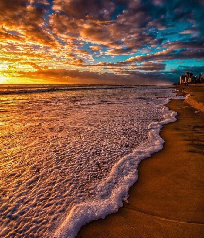 the sun is setting over the ocean with waves coming in to shore and buildings on the horizon