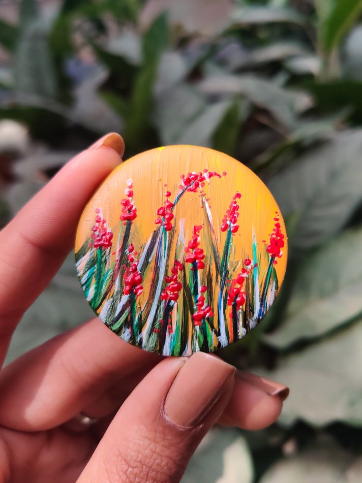 a hand holding a yellow button with red flowers on it in front of green plants