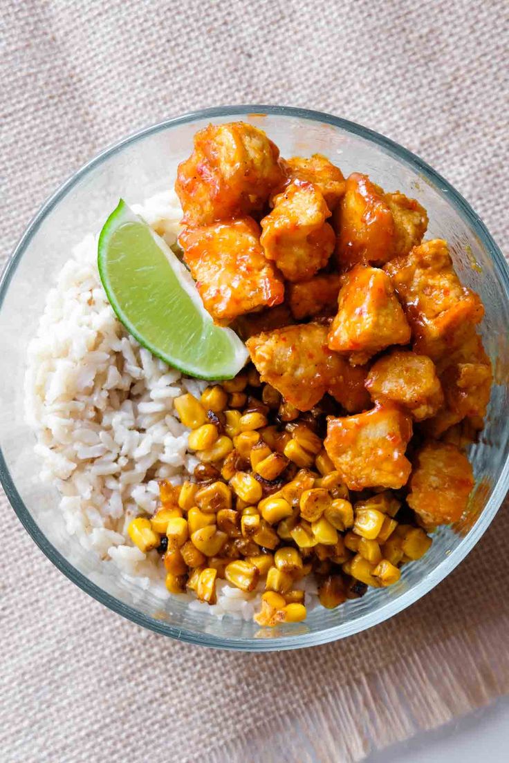 a glass bowl filled with rice, corn and tofu next to a lime wedge