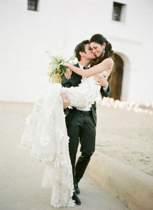 a bride and groom kissing in front of a white building with flowers on the bouquet