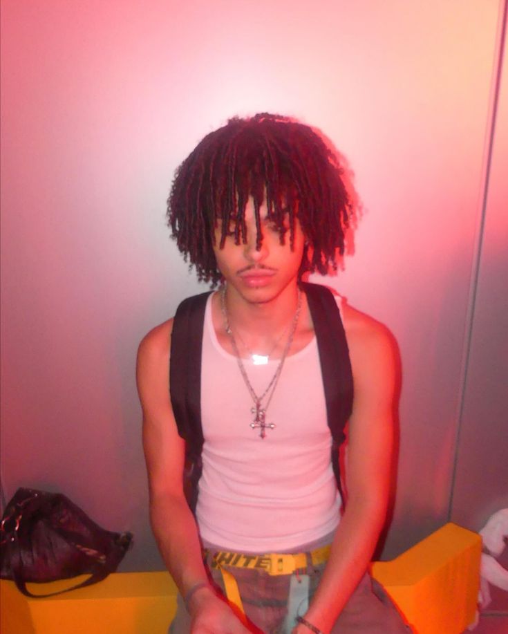 a young man with dreadlocks is sitting on a yellow bench in front of a white wall