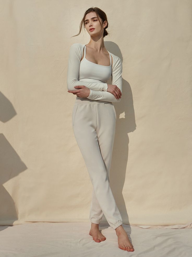 a woman standing in front of a beige wall wearing white pants and a long sleeved top