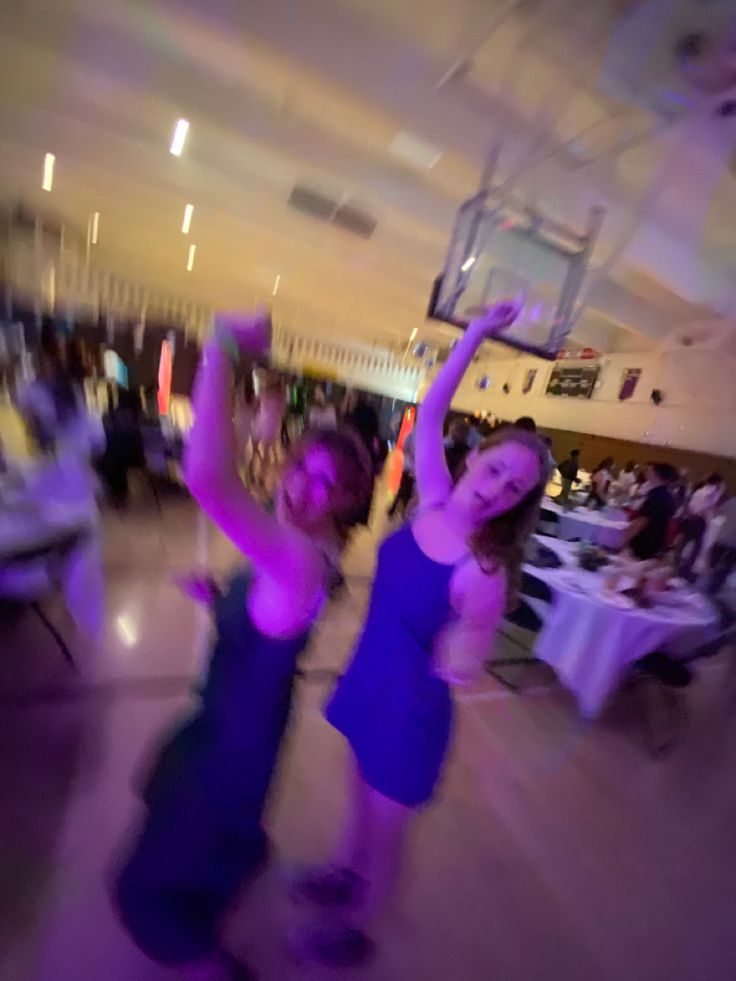 two women dancing at a party with purple lights on the dance floor and people seated in the background