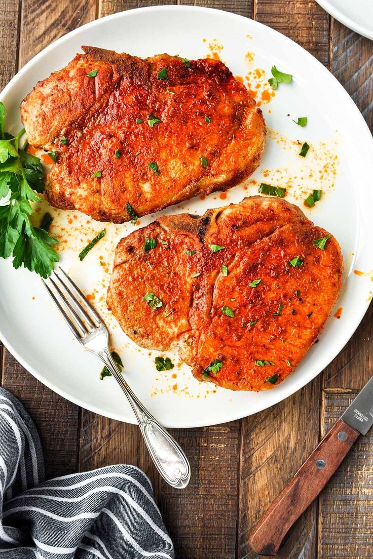 two pork chops on a white plate with parsley next to the knife and fork