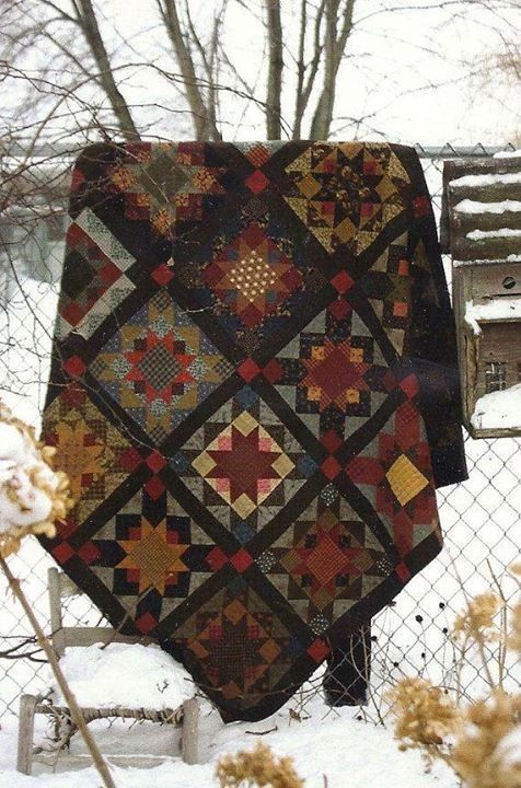 a quilt is hanging on a fence in the snow, with some flowers behind it