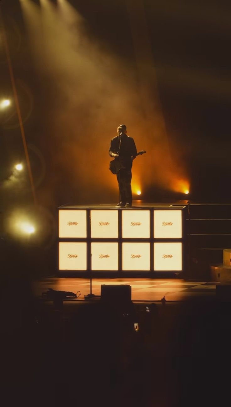 a man standing on top of a stage holding a guitar in front of two spotlights