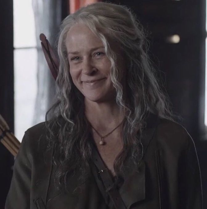 a woman with long gray hair and an arrow in her hand smiles at the camera