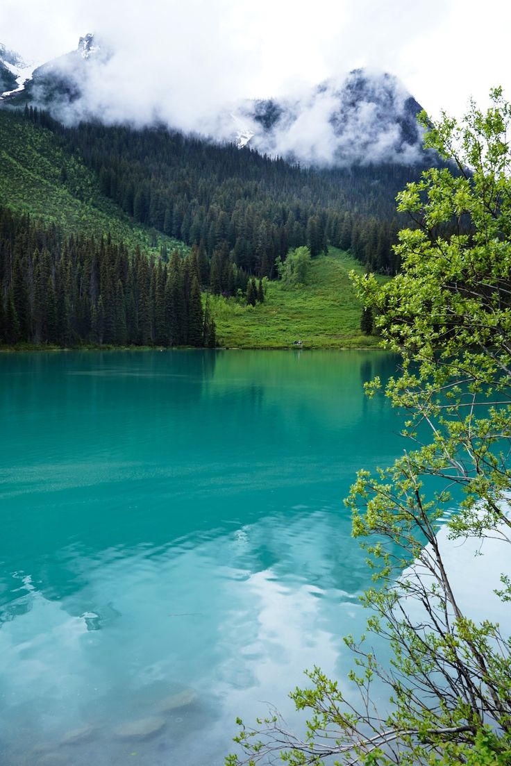 a lake surrounded by trees and mountains in the distance with clouds hanging over it's sides