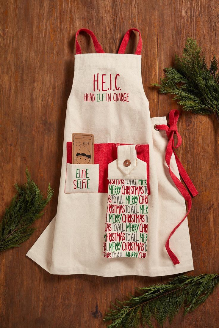 a white apron with red and green designs on it sitting on a wooden table next to evergreen branches