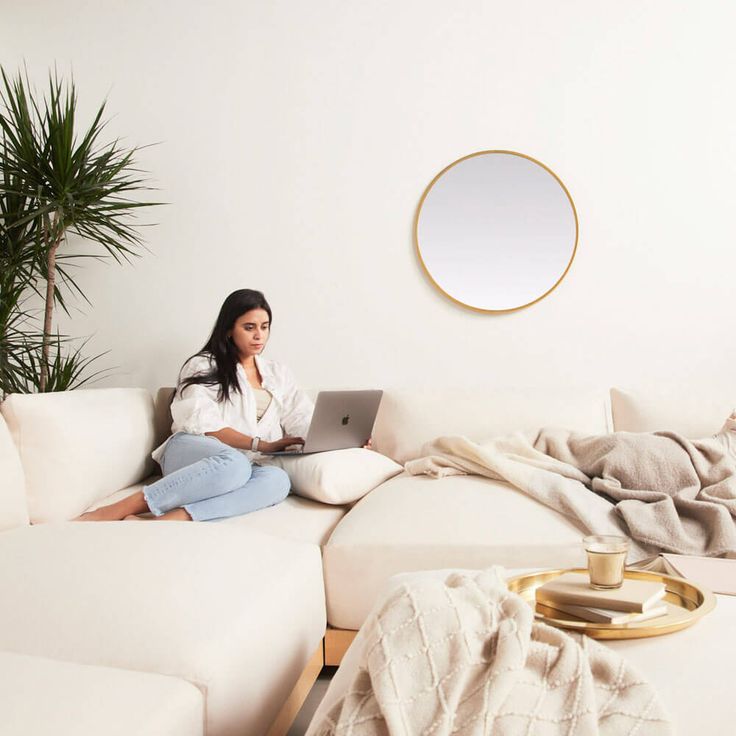 a woman sitting on top of a white couch in front of a laptop next to a potted plant