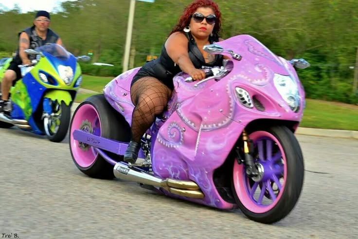 two people riding on the back of a pink motorcycle down a street next to trees