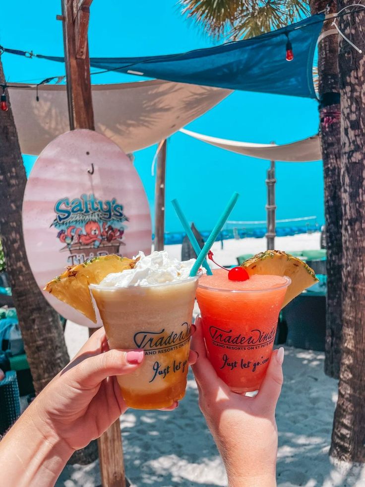 two people holding up drinks in front of palm trees on the beach with blue sky