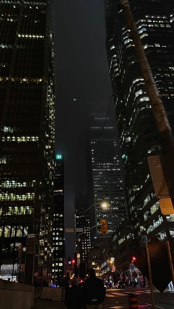 two people are walking down the street in front of some tall buildings at night time