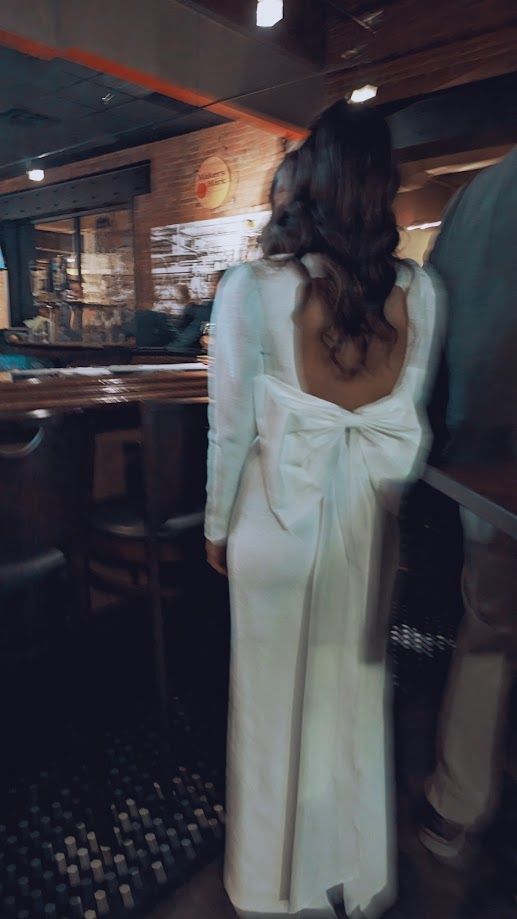 a woman standing in front of a bar wearing a white dress with a bow on the back