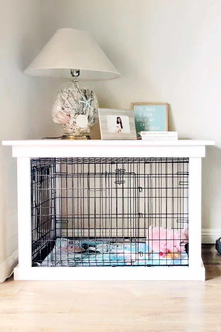 a dog cage sitting on top of a white table next to a lamp and pictures