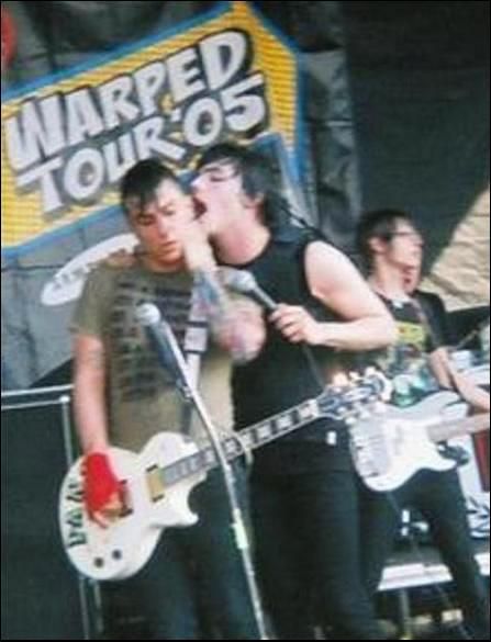 two young men playing guitars on stage at an outdoor music festival, one man is singing into the microphone