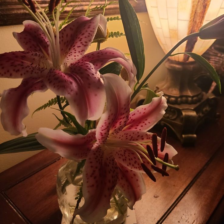 a vase filled with pink lilies on top of a wooden table next to a lamp