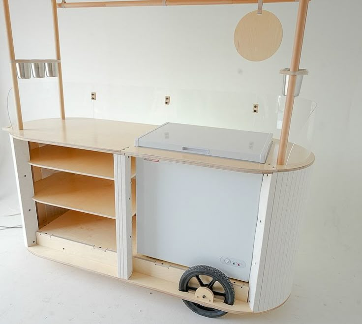 a small wooden cart with wheels and shelves on the side, in front of a white wall