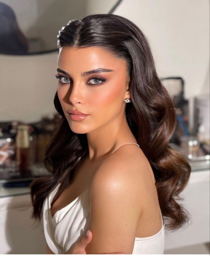 a woman with long dark hair wearing a white dress and pearls in her hair is looking at the camera