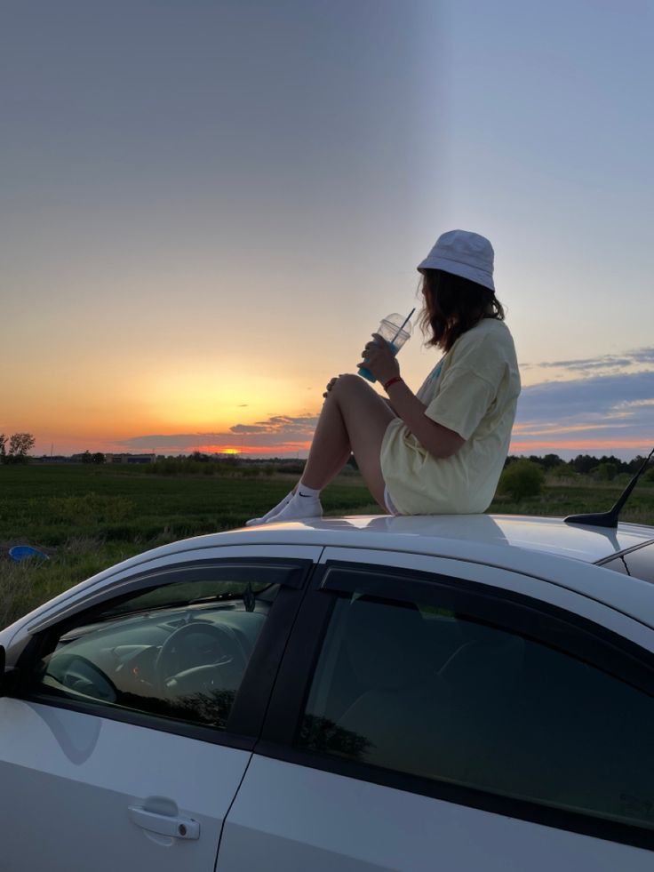 a woman sitting on top of a white car drinking from a wine glass at sunset