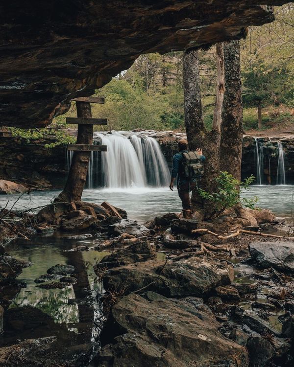 a man standing in front of a waterfall under a large rock formation next to a forest