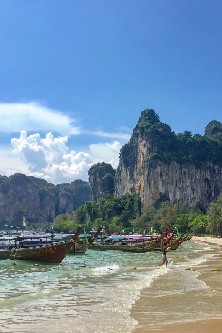 several boats on the beach with mountains in the background and people walking around them near the water