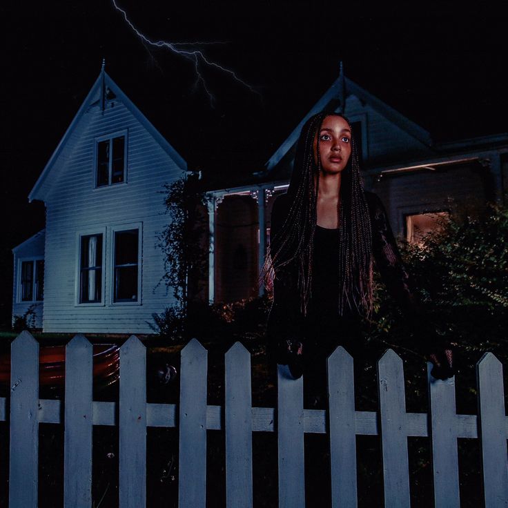 a woman standing in front of a white picket fence at night with a lightning behind her