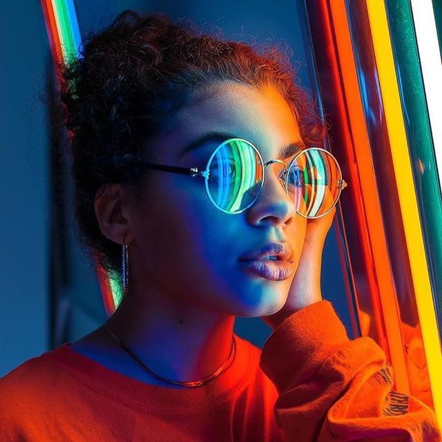 a young woman wearing glasses and standing in front of neon colored poles with her hand on her head