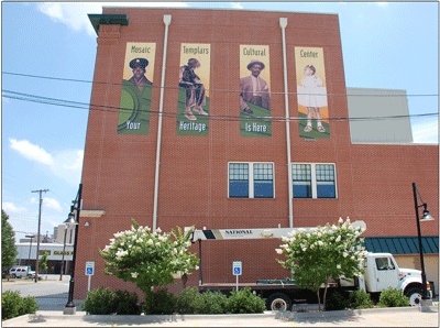 a large brick building with three murals on the side and one has a white truck parked in front of it