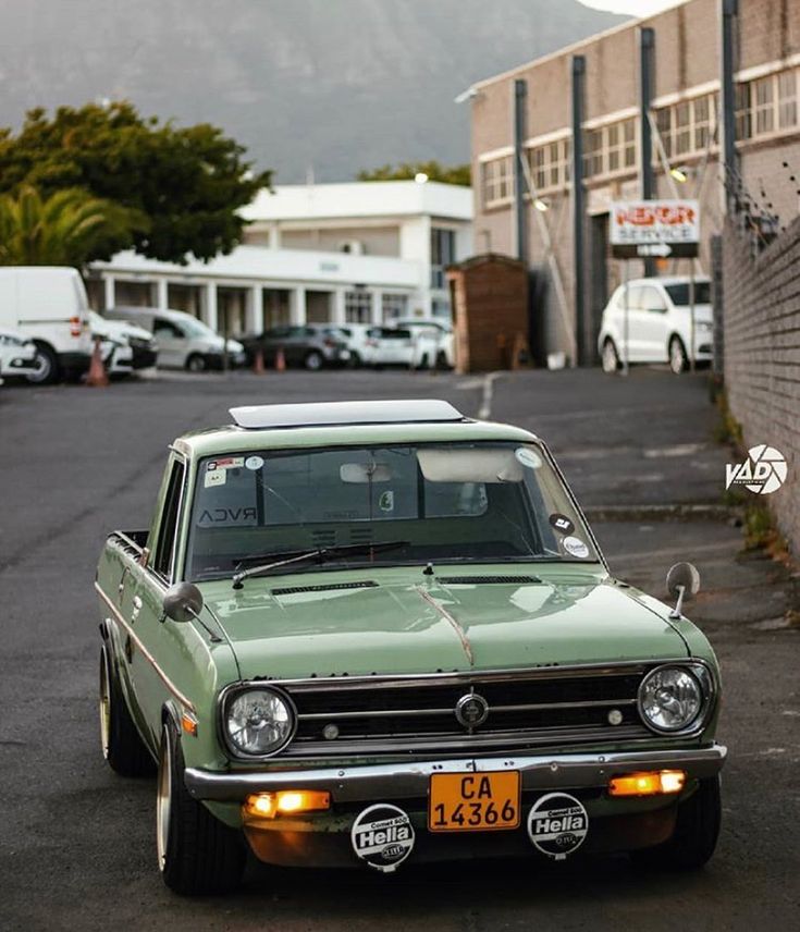 an old green car parked in front of a building with mountains in the back ground