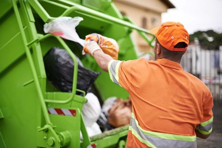 a man in an orange safety vest is dumping garbage into a green dumpster truck