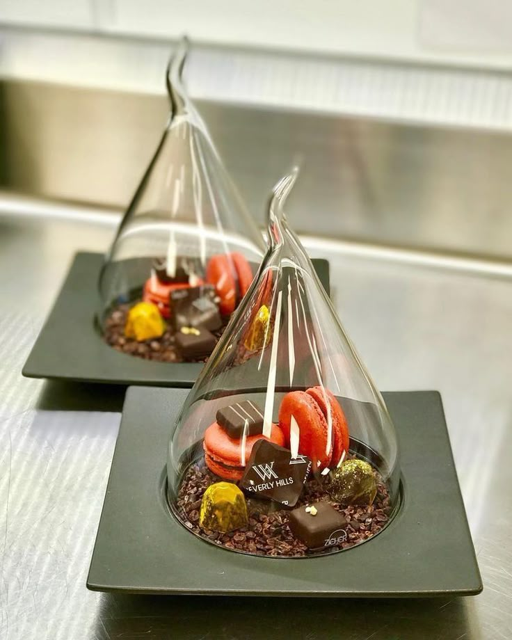 two chocolate covered desserts are sitting on black trays, one is shaped like a cone and the other has strawberries in it