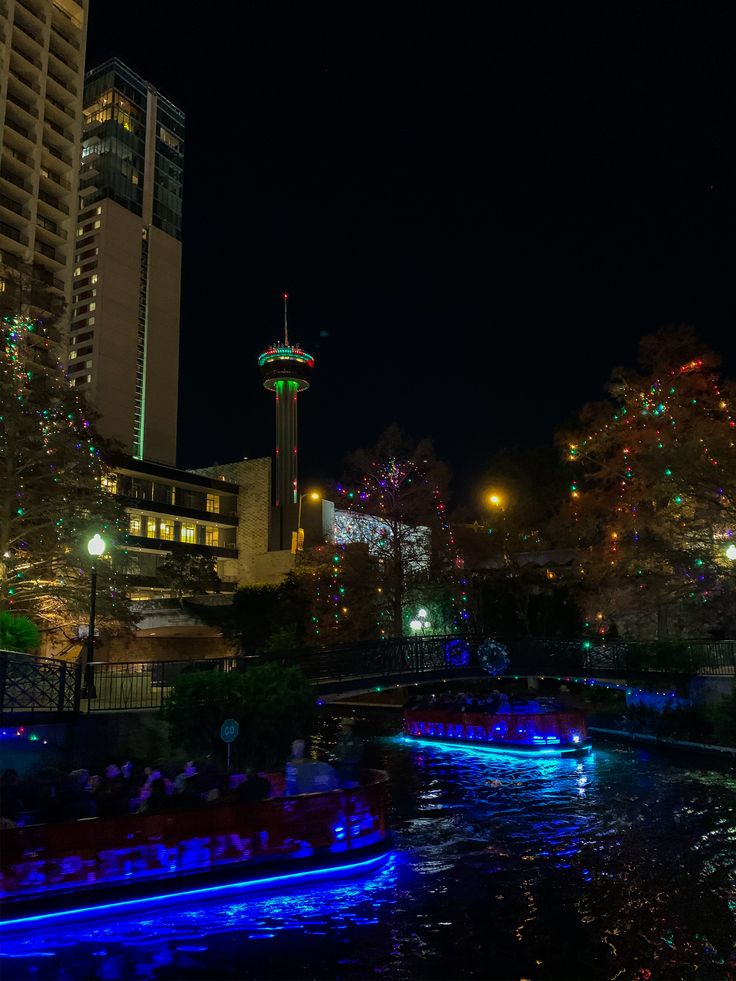 boats float down the river at night with christmas lights in the trees and buildings behind them