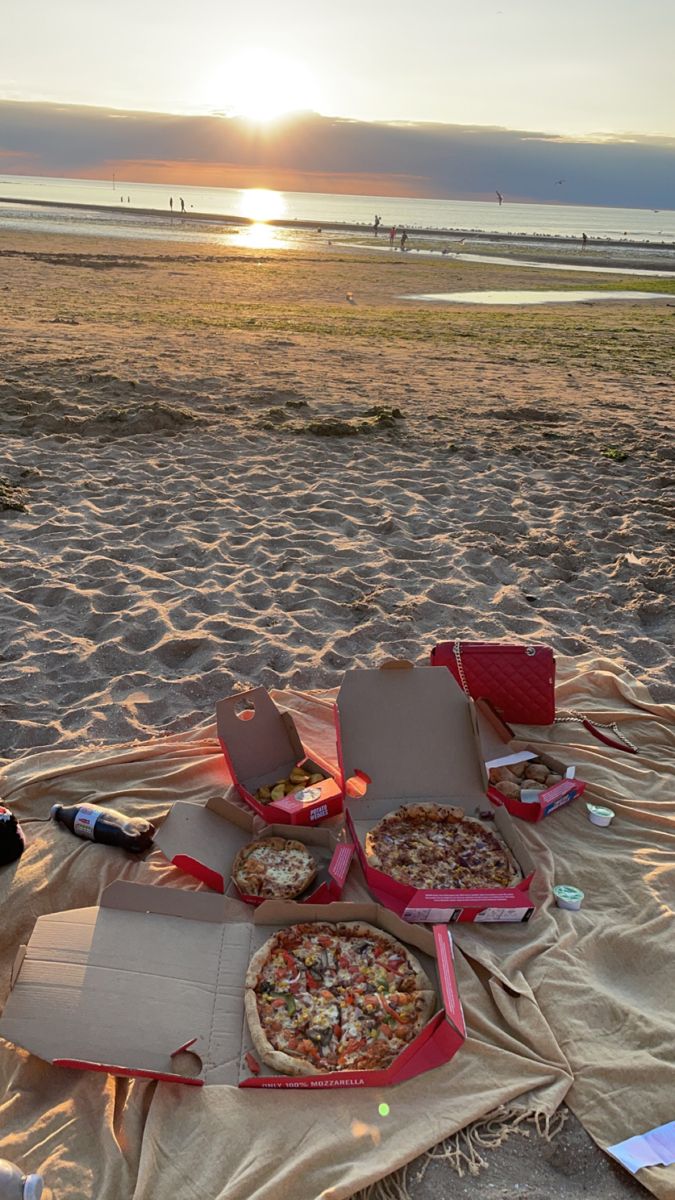 several pizzas sitting on top of a blanket on the beach near the ocean at sunset